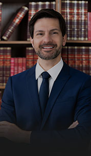 Homem branco sorrindo de roupa social em frente a uma estante com livros de direito